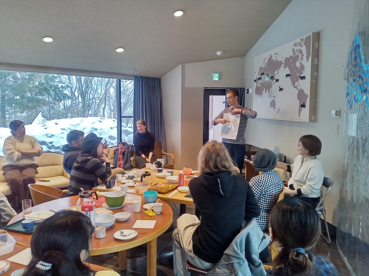 A group of people gathered in a room with large windows showing a snow-covered landscape outside. A person wearing a striped shirt, Peter Orosz, is standing and presenting something while holding a white sheet. The audience is seated around wooden tables covered with dishes, cups, and snacks, attentively watching the presenter. On the wall behind the presenter is a world map with small labels. The room has recessed lighting in the ceiling and blue curtains framing the window.