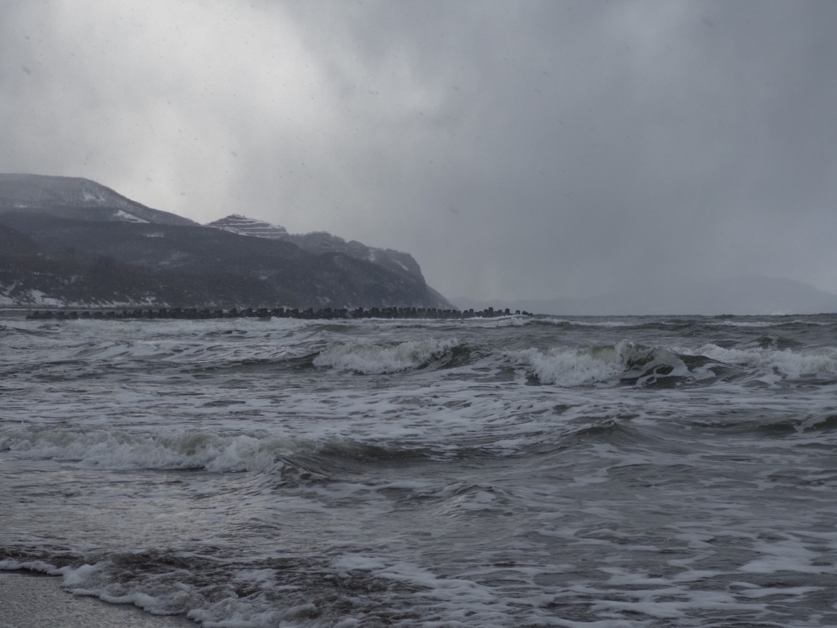 This image shows a dramatic winter seascape along the coast of the Sea of Japan, captured between Sapporo and Otaru. Choppy gray waves with white foam roll toward the shore in the foreground. In the background, snow-dusted mountains rise majestically, partially shrouded by heavy storm clouds. The scene has a moody, atmospheric quality with an overcast sky creating a monochromatic palette of grays and whites. A line of what appears to be coastal structures or breakwaters is visible along the shoreline. The overall impression is one of winter's stark beauty and the raw power of nature where sea meets mountainous landscape.
