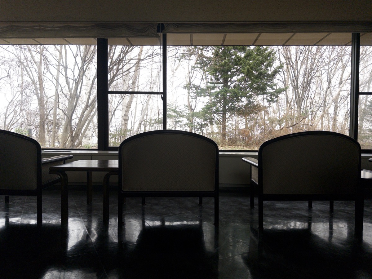 A row of four empty chairs sits silhouetted against large windows that stretch across the wall at Tenjinyama Art Studio in Sapporo, Hokkaido, Japan. The minimalist seating area features black-backed chairs with armrests arranged along a narrow counter that runs beneath the windows. Through the windows, bare winter trees and evergreens are visible against a gray sky, creating a stark contrast with the dark interior of the studio space. The polished floor reflects the natural light coming through the windows.